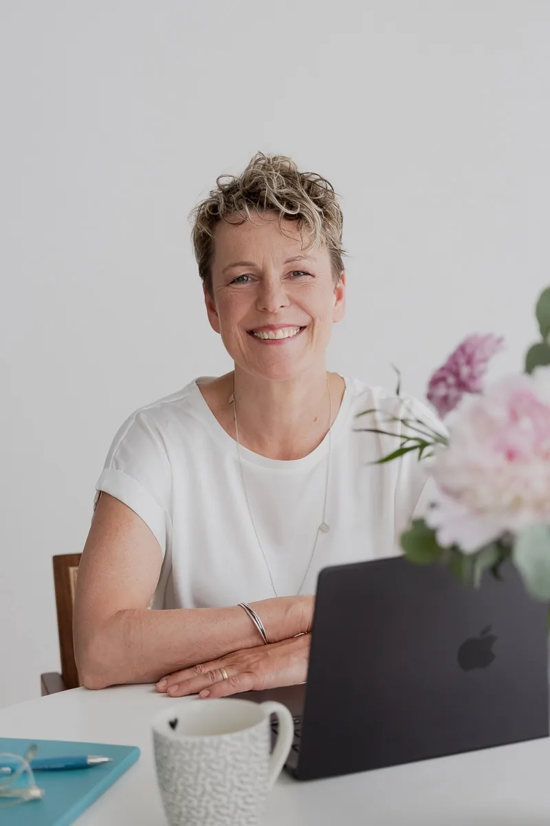 Britta Wein sitzt an einem Tisch mit Laptop und Blumen und blickt in die Kamera.