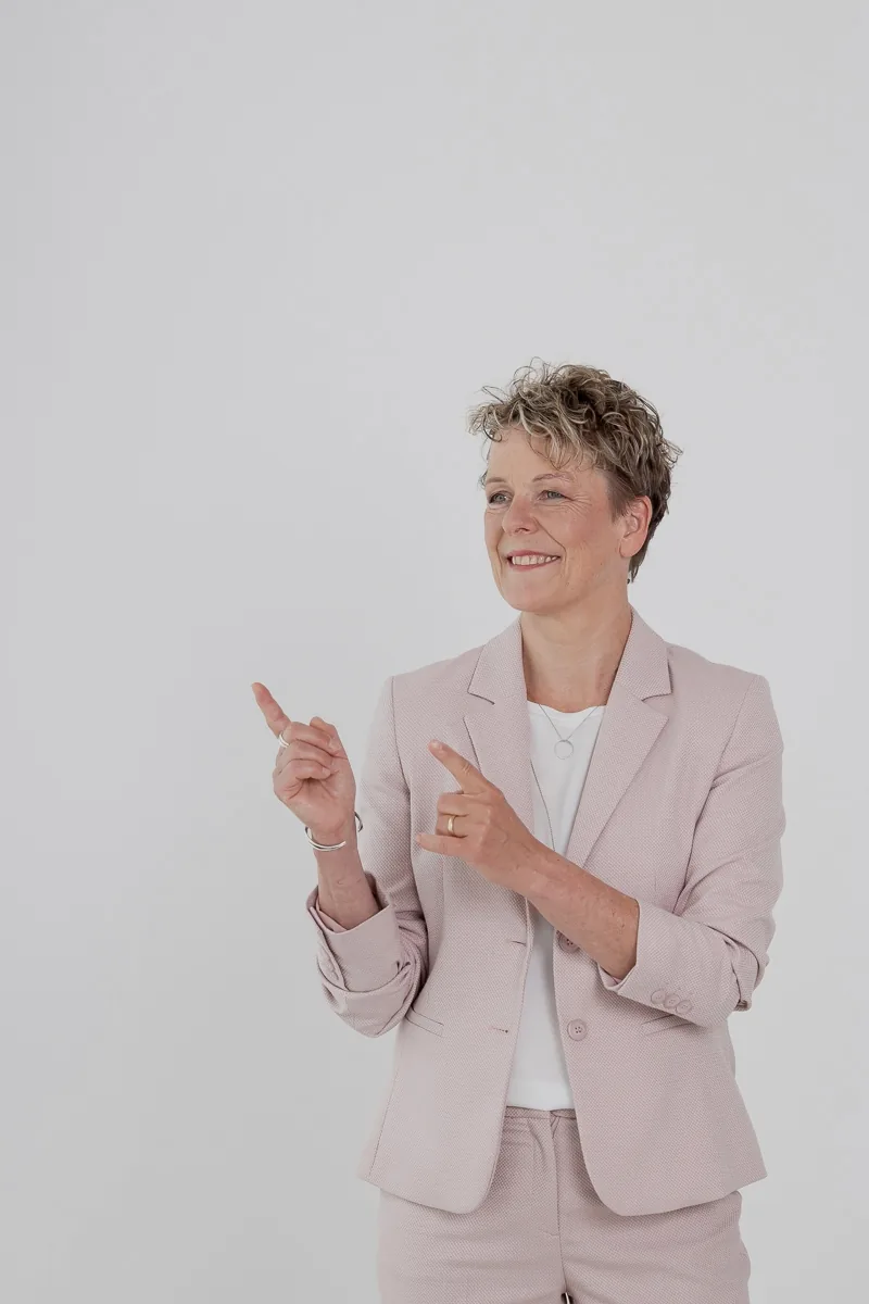 Britta Wein steht im rosafarbenen Anzug und zeigt mit beiden Händen nach links.